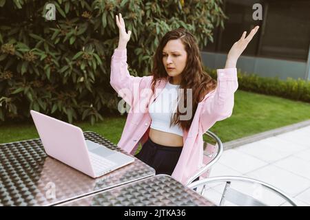 Junge Frau in legerer Kleidung mit Laptop wütend, verärgert und enttäuscht, heben ihre Hände mit Überraschung, sitzen im Straßencafe. Freiberuflich, Online-Geschäft, Online-Arbeit. Speicherplatz kopieren Stockfoto