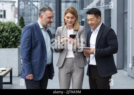 Drei UNTERNEHMER AUS DEM IT-Bereich vor dem Bürogebäude sehen sich am Telefon eines Kollegen ein Video an, glücklich und lächelnd, während sie sich in der Pause von der Arbeit ausruhen Stockfoto