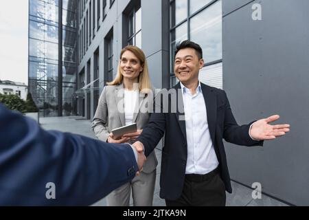 Männer in Geschäftsanzügen grüßen und schütteln die Hände, ein Team aus verschiedenen Geschäftsleuten trifft sich vor dem Bürogebäude Stockfoto