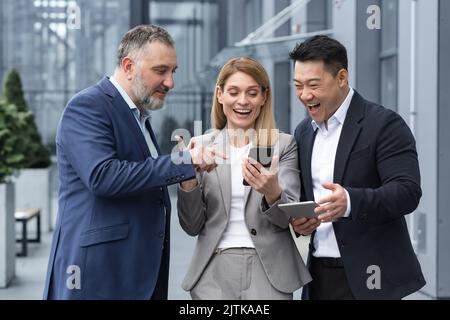 Drei UNTERNEHMER AUS DEM IT-Bereich vor dem Bürogebäude sehen sich am Telefon eines Kollegen ein Video an, glücklich und lächelnd, während sie sich in der Pause von der Arbeit ausruhen Stockfoto