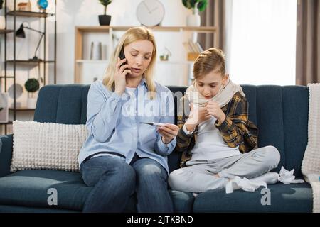 Behandlung der Grippe zu Hause. Mutter hält Thermometer und Handy, ruft den Arzt an, während sie die Temperatur ihres kranken Teenagersohnes überprüft, der mit hohem Fieber hustet und auf dem Sofa sitzt. Stockfoto