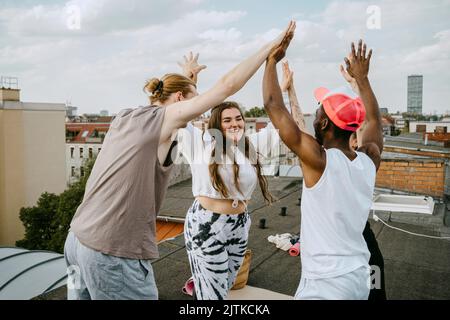 Glückliche multirassische männliche und weibliche Freunde mit aufgehobenen Armen, die sich gegenseitig auf dem Dach hoch-fünf geben Stockfoto