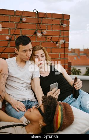 Junge Männer teilen sich Smartphone, während sie auf dem Dach sitzen Stockfoto