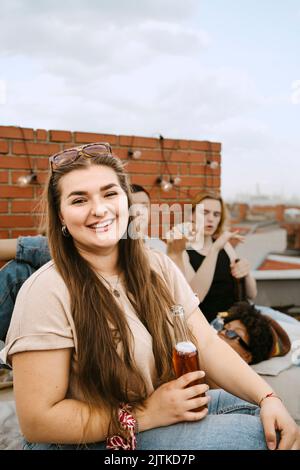 Porträt einer lächelnden jungen Frau, die eine Bierflasche in der Hand hält und vor Freunden auf dem Dach sitzt Stockfoto