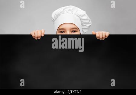 Kleiner Junge in der Haube des Küchenchefs hinter schwarzer Tafel Stockfoto