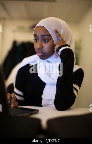 Nachdenkliches Mädchen trägt Schal mit Laptop im Klassenzimmer Stockfoto