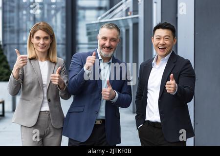 Fröhliches Dream Team, erfahrene und ERFAHRENE IT-Profis in Geschäftsanzügen lächelnd und glücklich, den Daumen hoch haltend und auf die Kamera schauend, vielfältige Gruppe von Projektmanagern außerhalb des Bürogebäudes. Stockfoto