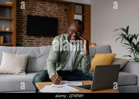 Ein fröhlicher afroamerikanischer Mann mit Brille arbeitet am Laptop mit Dokumenten und telefoniert im Wohnzimmer Stockfoto