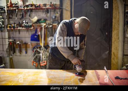 Mann grinst den Tisch. Mann in der Werkstatt arbeitet an Bord. Mann und viele Werkzeuge. Arbeiter hält Elektrowerkzeug. Stockfoto