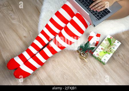 Mädchen in Weihnachtskniesocken sitzen auf einem Pelzteppich mit Laptop in der Nähe der Geschenkboxen. Bestellung von Geschenken, Arbeit und Freizeit zu Hause während der Neujahrsferien Stockfoto