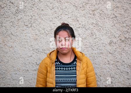 Viacha, La Paz, Bolivien - 16 2022. August: Eine bolivianische Frau in einer gelben Winterjacke blickt auf die Kamera und posiert vor einer strukturierten Wand Stockfoto