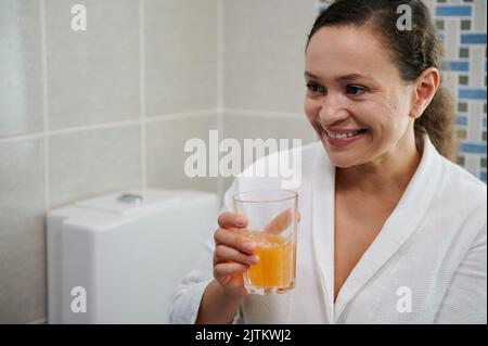 Frau mittleren Alters im Bademantel hält ein Glas Wasser mit Brausetablette, nimmt tägliche Dosis von Vitaminen und Mineralstoffen Stockfoto