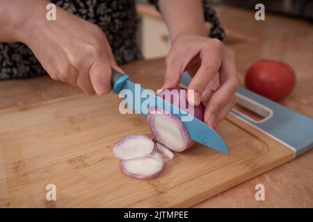 Nahaufnahme von Frauenhänden, die eine Zwiebel mit einem blauen Messer auf einem Holzbrett an der Küchentheke schneiden Stockfoto