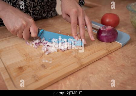 Nahaufnahme der Hände einer Frau, die Zwiebeln mit einem blauen Messer auf einem Holzbrett an der Küchentheke hackt Stockfoto