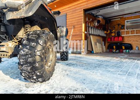 Nahaufnahme des hinteren POV-Nahaufnahme des Quad-Bike-Offroad-Fahrzeugs, das an einem sonnigen, schneebedeckten, kalten Wintertag in der Garage vor dem Haus geparkt ist. ATV-Abenteuer extrem Stockfoto