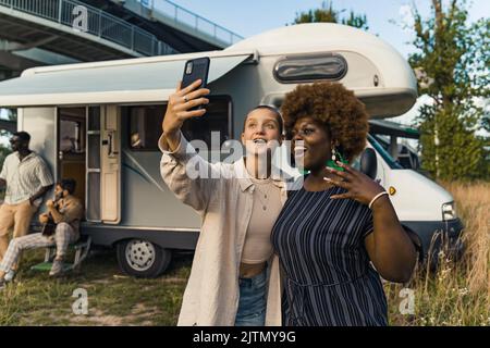 Interrassische Freundschaft. Zwei junge, vielfältige Mädchen, die in der Nähe ihres Wohnwagens ein Selfie am Telefon machen. Die Jungs sitzen auf der Treppe und spielen im Hintergrund Gitarre. Wieder vereint auf ihrem Camping-Van-Trip durch das Land. Hochwertige Fotos Stockfoto