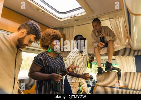 Interracial Gruppe von engen Freunden tanzen zur Musik in ihrem komfortablen geräumigen Wohnmobil und trinken Bier. Wir feiern im Sommer einen Geburtstag auf einer Campingbus-Reise. Hochwertige Fotos Stockfoto