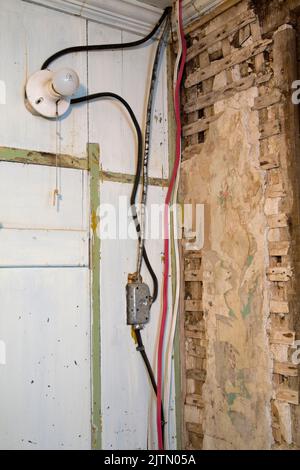 Alte Elektroblechisolierung und Kunststoffverkabelung mit Glühbirnenhalterung und Steckdose auf weißem Putz und Holzgitterwand in einem leeren Raum. Stockfoto