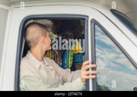 Junge kaukasische hübsche Frau mit einem rasierten Kopf sitzt neben dem offenen Fenster eines Campingwagens und diskutiert etwas mit ihrer afrikanischen Freundin auf dem Rücksitz. Hochwertige Fotos Stockfoto