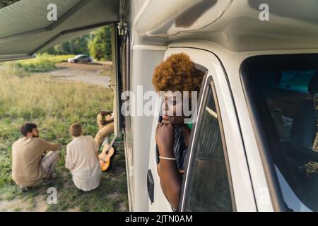 Nachdenkliche dunkelhäutige afrikanische junge Frau mit verworrem Haar, das aus dem Fenster eines Wohnwagens gelehnt ist, und ihre Freunde, die am Ende ihrer Reise auf dem Gras neben ihr sitzen. Hochwertige Fotos Stockfoto