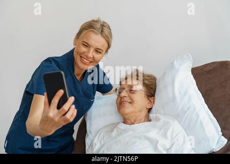 Arzt oder Krankenschwester, die sich um eine ältere Frau mit einem Mobiltelefon kümmert, nimmt ein Selfie im Krankenhauszimmer auf Stockfoto