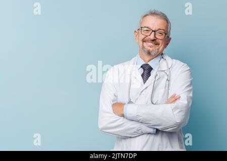 Lächelnder Arzt im Labormantel mit gekreuzten Armen vor blauem Hintergrund. Stockfoto