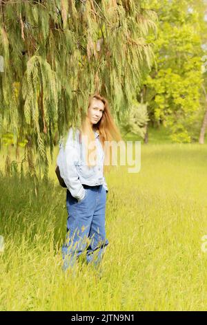 Frau auf einer Wiese unter Ästen eines Baumes. Frau im Park unter einem Thuja-Baum Stockfoto