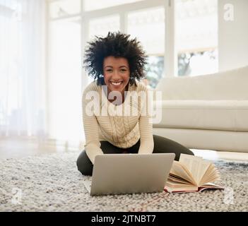 Ich recherchiere für meinen Buchclub. Porträt einer attraktiven jungen Person, die ihren Laptop benutzt und ein Buch liest, während sie zu Hause auf dem Boden sitzt. Stockfoto
