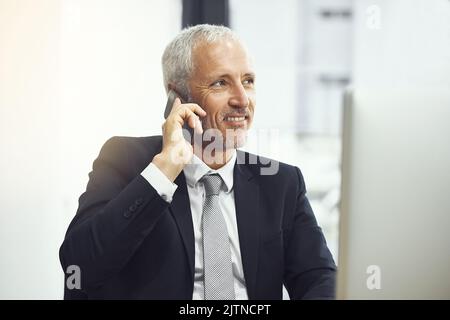 Ein lächelnder, reifer Geschäftsmann, der am Telefon telefoniert, während er in einem Büro arbeitet. Stockfoto