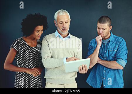 Wenn Sie hier genau hinschauen... ein reifer Geschäftsmann, der zwei jüngeren Kollegen im Studio etwas auf einem Tablet zeigt. Stockfoto