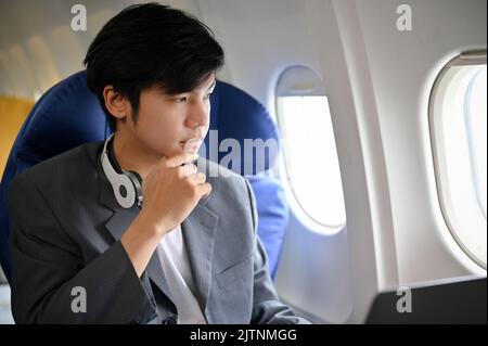 Ein eleganter und hübscher junger asiatischer Geschäftsmann im formellen Anzug ist auf dem Flug, sitzt in der Business-Klasse, blickt aus dem Flugzeugfenster und denkt nachdenklich an Stockfoto