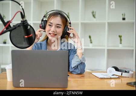 Schöne attraktive junge asiatische weibliche Radio-Host oder Podcasterin trägt Kopfhörer, sprechen auf ihrem professionellen Mikrofon, läuft ihr Podcast chann Stockfoto