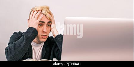 Der junge Büroangestellte ist schockiert und überrascht, als er einen Laptop sieht, der seinen Kopf umklammert und an einem Schreibtisch in einem Büro sitzt, das von Dokumenten und Notebooks umgeben ist Stockfoto