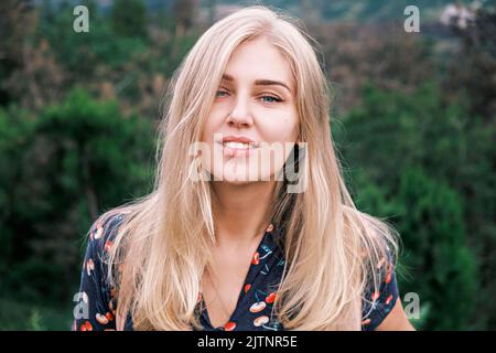 Porträt einer jungen attraktiven blonden Frau mit blauen Augen, langen Haaren, die in die Kamera schaut und vor einem verschwommenen Hintergrund im Freien lächelt Stockfoto