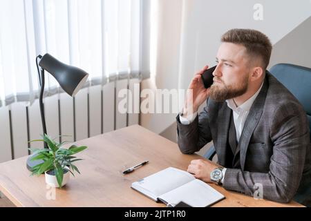 Der bärtige Mann aus der Geschäftswelt telefoniert und plant, sich täglich zu treffen und eine Geschäftsreise in ein Tagebuch am Schreibtisch zu schreiben. Kalender-Planer und Agenda-Erinnerungen. Stockfoto