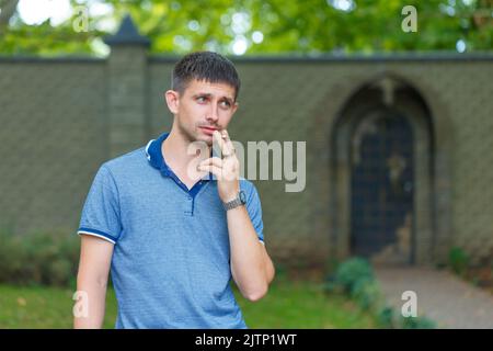 Ein unrasierter Mann steht auf der Straße und raucht in Erwartung von Gedanken und Erfahrungen, tagsüber in einem blauen T-Shirt und Jeans vor dem Hintergrund eines Steinzauns Stockfoto