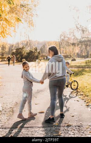 Rückansicht einer Mutter, die morgens mit ihrer Tochter im Park unterwegs war, das Mädchen drehte ihren Kopf und lächelte die Kamera an. Sommer Stockfoto