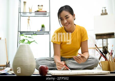 Attraktive junge asiatische Studentin, die einen Kunstunterricht im Kunststudio hat, Zeichnen und Malen auf Leinwand übt, lächelt und schaut auf die Kamera. A Stockfoto