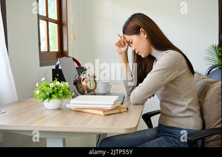 Junge asiatische College-Studentin oder Freiberuflerin, nachdenklich und gestresst, hart an ihrem Schreibtisch arbeitend, über ihren Arbeitsauftrag nachdenkend und planend. Stockfoto