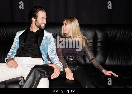 Fröhliches, stilvolles Paar, das sich anschaut, während es auf der Ledercouch auf Schwarz sitzt Stockfoto