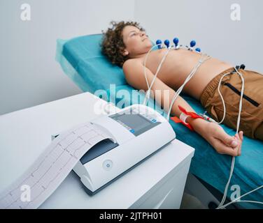 EKG-Schreiber mit EKG-Ausdruck für einen Teenager-Jungen, der im medizinischen Bett mit Vakuumsensoren liegt. Herzelektrokardiographie für Kinder Stockfoto