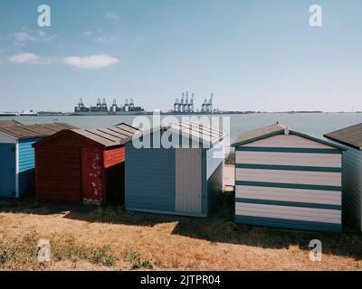 Bemalte Strandhütten an der Strandpromenade in Harwich mit Blick auf den Hafen von Felixstowe an der Küste von Essex im Sommer in England - Containerhafenkrane Stockfoto