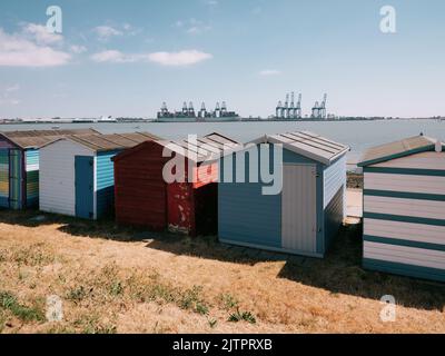 Bemalte Strandhütten an der Strandpromenade in Harwich mit Blick auf den Hafen von Felixstowe an der Küste von Essex im Sommer in England - Containerhafenkrane Stockfoto