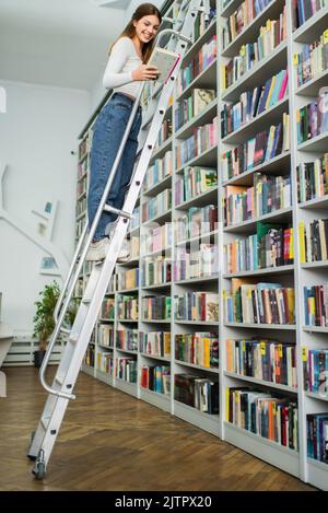 Glückliches Teenager-Mädchen hält Buch, während auf Leiter in der Bibliothek stehen, Stock Bild Stockfoto