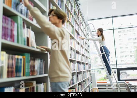 Teenager-Mädchen auf Leiter stehen und wählen Bücher in der Nähe Freund auf verschwommenem Vordergrund, Stockbild Stockfoto