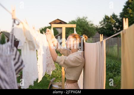 Wäschereitag. Eine Frau hängt Bettwäsche und Handtücher auf einem Baum im Hof eines Dorfhauses. Sommerhaus-Konzept. Stockfoto