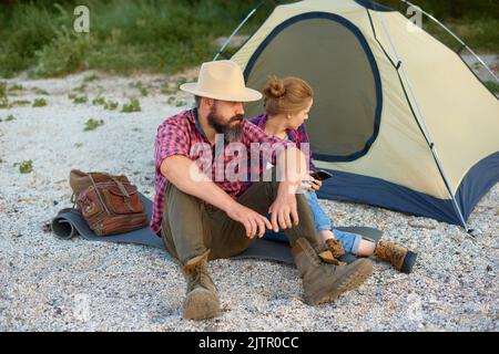Pärchen, die sich am Strand ausruhen. Weiße Freunde sitzen am Herbsttag beim gemeinsamen Campen in der Nähe des Zeltes Stockfoto