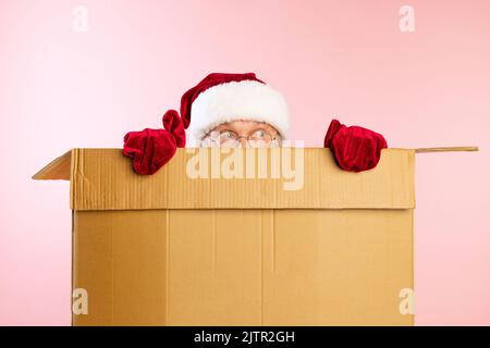 Porträt eines älteren Mannes im Bild des Weihnachtsmanns, der sich in einem Karton versteckt. Geschenke, Geschenke für den Urlaub Stockfoto