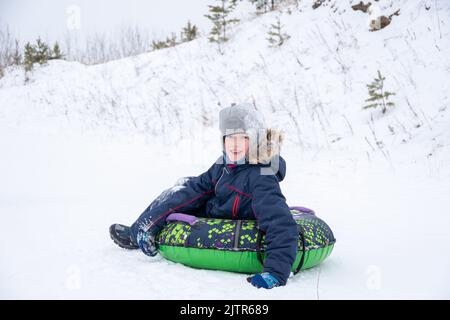 Glücklich lächelnder kleiner Junge rutscht die Eisrutsche hinunter, sitzt auf einem aufblasbaren Schlauch. Nettes kleines glückliches Kind, das im Winter auf Schlitten Spaß im Freien hat. famii Stockfoto