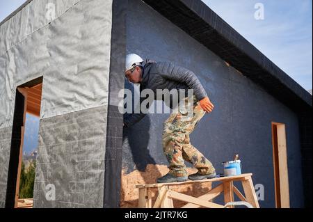 Männlicher Maler mit Farbrolle, tun Außenlackarbeiten in einer schwarzen Farbe. Mann Arbeiter Gebäude Holzrahmen Haus. Zimmerei- und Konstruktionskonzept. Stockfoto